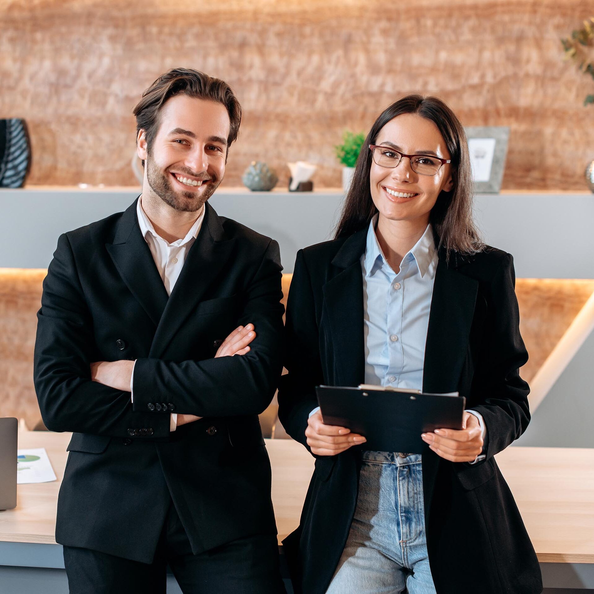 Portrait of two caucasian confident successful company employees, business colleagues, a man and a woman, stand near the desktop in a modern office, look at the camera, smile friendly